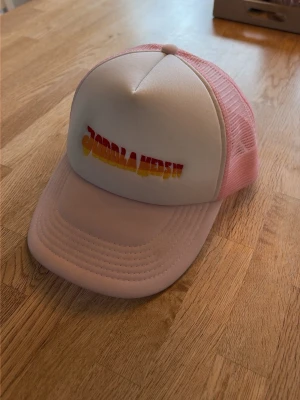 Rosa joddla med Siv keps - Säljer en snygg joddla med Siv truckerkeps i rosa och vitt med färgglad text framtill. Kepsen har böjd skärm, mesh på sidorna och justerbar snapback. Perfekt för en cool och avslappnad stil.