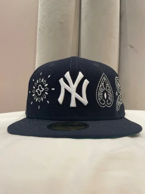 Mörkblå fitted New York Yankees keps - Snygg mörkblå 59FIFTY keps från New Era med New York Yankees-logga i vitt framtill. Kepsen har broderade paisleymönster och detaljer i vitt på framsidan och sidan. Klassisk rak skärm. Perfekt för dig som gillar streetwear.