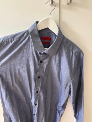 Blå skjorta från Hugo Boss - Snygg blå skjorta från Hugo Boss med klassisk krage och svarta knappar. Skjortan är tillverkad i ett super skönt och tunt material! 