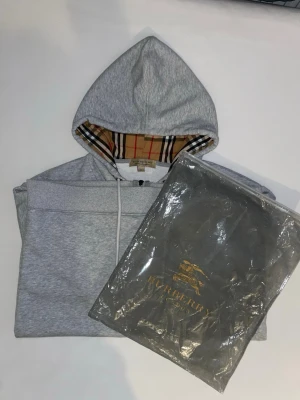 Grå zip hoodie från Burberry - Snygg grå zip hoodie från Burberry med klassiskt rutigt foder i huvan och diskret logga på bröstet. Hoodien har dragsko i huvan, fickor framtill och ribbade muddar. Perfekt för dig som vill ha en stilren och exklusiv look.