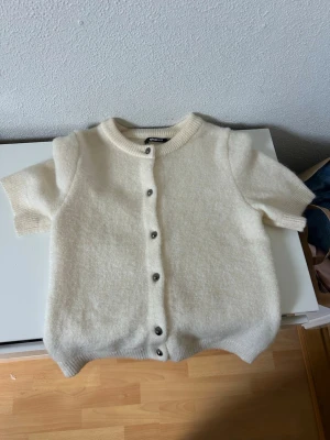 Krämvit kortärmad kofta - Mysig krämvit kofta med korta ärmar och rund hals. Koftan har ribbstickade kanter och knäpps med silvriga knappar framtill. Perfekt att slänga över en topp för en enkel men stilren look. Använd fåtal gånger 