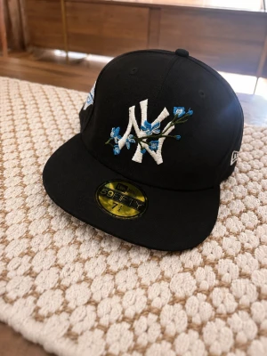 Svart New Era NY keps med blommor - Snygg svart 59FIFTY keps från New Era med New York Yankees-logga framtill, dekorerad med blå broderade blommor. Platt skärm med ljusblå undersida och broderad baseboll. Klassisk snapback-stil och broderade detaljer på sidorna.