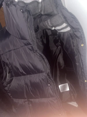 Svart dunväst från burbery  - Snygg svart dunväst med quiltad design och hög krage. Västen har dragkedja framtill och tryckknappar, samt två sidofickor. Perfekt för lager-på-lager och håller dig varm under kyliga dagar. Materialet är glansigt och ger en modern look. Knappt använd heller 