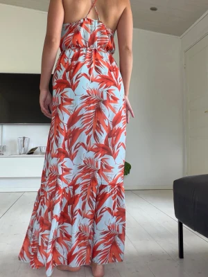 Lång sommarklänning med palmbladsmönster - Snygg långklänning med tunna axelband och v-ringning. Klänningen har ett tropiskt palmbladsmönster i rött och orange på ljusblå botten. Resår i midjan för skön passform och luftig känsla. Perfekt för varma dagar och semester.