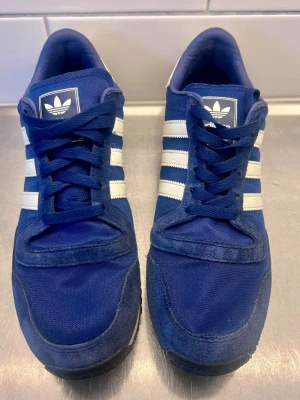 Blå Adidas sneakers med vita ränder - Snygga blå sneakers från Adidas med klassiska vita ränder på sidorna. Ovandelen är i en mix av mocka och textil, och skorna har blå snörning samt svart yttersula. Perfekta för en sportig och avslappnad stil.