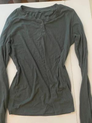 Mörkgrön ribbad långärmad topp - Snygg mörkgrön ribbad topp med lång ärm och två knappar framtill. Perfekt till jeans eller kjol för en enkel men trendig look. Materialet känns mjukt och stretchigt, vilket gör den bekväm att bära hela dagen.