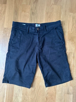 Svarta shorts från Jack & Jones strl S - Snygga svarta shorts i bomull med klassisk passform och knäppning fram. De har sidofickor och två bakfickor med minimalistisk design. Perfekta för varma dagar och enkla att matcha med olika stilar.