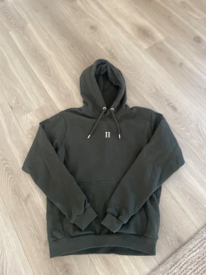 Mörkgrön hoodie från Les Deux - Snygg mörkgrön hoodie från Les Deux med broderad II-logga på bröstet. Klassisk känguruficka framtill, ribbade muddar och justerbar huva med snören. Tillverkad i mjukt bomullsmaterial som känns skönt mot huden