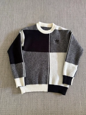 Blockmönstrad stickad tröja Adidas - Säljer en stickad tröja från Adidas med blockmönster i svart, grått och vitt. Tröjan har rund halsringning, ribbade muddar och Adidas-logga broderad på bröstet. Perfekt för dig som gillar streetwear och grafiska mönster.