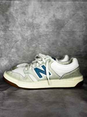 Vita New Balance sneakers med blå detalj - Snygga vita sneakers från New Balance med blått N på sidan och ljusgrå mockadetaljer. Ovandelen är i mesh och mocka, sulan är vit med brun yttersula. Klassisk retrostil som passar till många outfits.