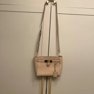 Michael Kors Hamilton Axelremsväska  - Beige axelväska med eleganta gulddetaljer. Färgen kan upplevas lite beigerosa. Väskan har en justerbar axelrem och ett dekorativt lås framtill. Överlag fint skick, det som finns att anmärka på syns på bild 3, mindre svart streck längst upp till höger samt i botten inuti väskan så är det en del smink rester som ev går att tvätta bort.