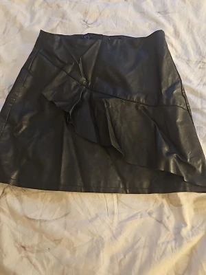 Svart kortkjol i skinnimitation - Snygg svart kortkjol i skinnimitation med volangdetalj framtill. Kjolen har en enkel, rak passform och stängs med dragkedja bak. Perfekt för dig som vill ha en edgy look med trendiga detaljer.