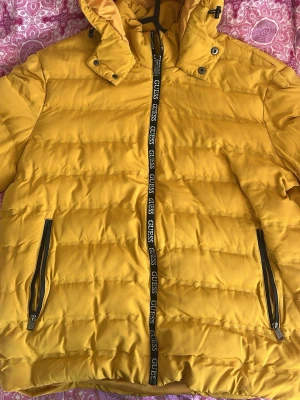 Gul pufferjacka från Guess - Snygg gul pufferjacka från Guess med huva och svarta dragkedjor. Jackan har Guess-logga längs dragkedjan framtill och två  fickor med dragkedja. Perfekt för kalla dagar och riktigt skön att bära. Materialet är quiltat och ger en modern look. Endast använd ett antal par gånger. Kan passa både XL och XXL