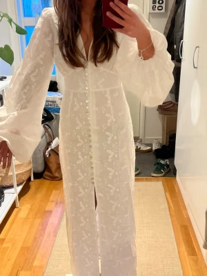 Adoore klänning med puffärmar - Elegant vit långklänning med puffärmar och blommigt broderi. Klänningen har v-ringning, knappar framtill och en slits nedtill. 