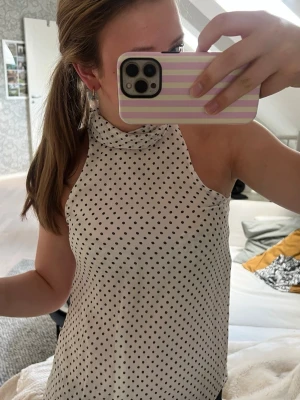 Prickig ärmlös blus med knyt - Snygg vit ärmlös blus med svarta prickar och hög hals. Blusen har en fin knytdetalj vid halsen som ger en trendig look. Säljer pga att den är för liten för mig, aldrig använt. Nypris 500
