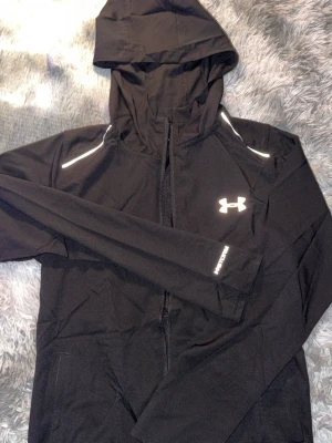 Svart vindjacka från Under Armour - Snygg svart vindjacka från Under Armour med huva och dragkedja framtill. Jackan har vita detaljer på axlarna och logga på bröstet samt stort Under Armour-tryck på ryggen. Perfekt för träning eller när du vill ha en sportig look.