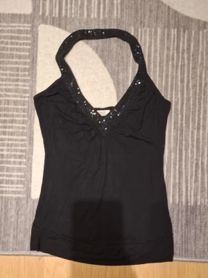 Svart paljettprydd halterneck topp - Snygg svart halterneck topp från ONLY med paljetter längs den djupa v-ringningen. Perfekt för dig som vill ha en enkel men ändå glittrig look. Toppen är ärmlös och har en smickrande passform.