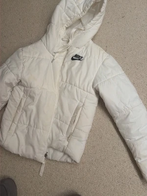 Vit pufferjacka från Nike med huva - Snygg vit pufferjacka från Nike med dragkedja framtill och två sidofickor. Jackan har en stor huva med dragsko och klassisk Nike-logga på bröstet. Perfekt för kalla vinterdagar och riktigt skön att bära.