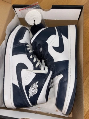 Air Jordan 1 Mid Obsidian/Vit - Snygga Air Jordan 1 Mid sneakers i färgerna obsidianblå och vit. Skorna har klassisk Nike Swoosh på sidorna, Air Jordan-logga vid ankeln och vita samt mörkblå skosnören. Tillverkade i läder med en bekväm och stabil sula. Perfekta för dig som gillar streetwear och ikonisk design.