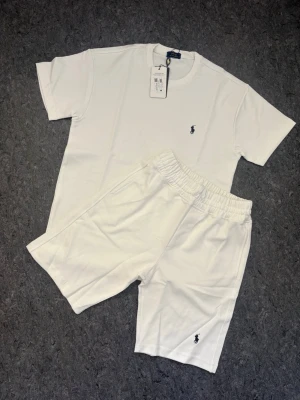 Ralph Lauren Set - Snyggt vitt set från Polo Ralph Lauren med t-shirt och shorts. Båda plaggen har den klassiska broderade loggan i svart. T-shirten har rund hals och kort ärm, shortsen har resår i midjan och dragsko. Perfekt för en avslappnad och stilren look.