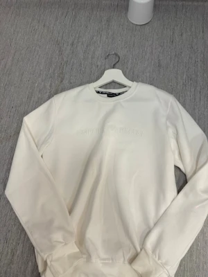 Vit sweatshirt från Emporio Armani - Snygg vit sweatshirt från Emporio Armani med diskret broderad logga framtill. Tröjan har rund halsringning, långa ärmar och ribbade muddar vid ärmslut och nederkant. Perfekt för en clean och stilren look.