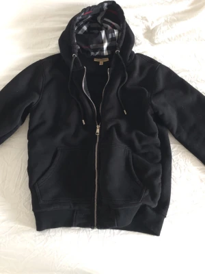 Svart hoodie med dragkedja från Burberry - Snygg svart hoodie från Burberry med dragkedja framtill och klassiskt rutigt foder i huvan. Storlek M men den passar bättre som S. Hoodien har fickor på magen, ribbade muddar och justerbar dragsko i huvan. Perfekt för en chill och stilren look. 