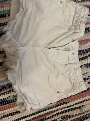 Vita fransiga jeansshorts - Säljer ett par vita jeansshorts med fransig kant och klassisk femficksmodell. Shortsen har hög midja, dragkedja och knapp framtill samt bälteshällor. Perfekta för sommaren och riktigt snygga till en enkel topp eller hoodie.