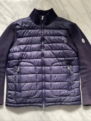 Marinblå Moncler cardigan utan luva  - Snygg mörklila cardiganjacka med quiltad framsida och stickade ärmar samt rygg. Jackan har hög krage, två framfickor med dragkedja och Moncler-logga på ena ärmen. Materialmix av vadderad nylon och stickad ull ger både värme och stil produkten är även lite solblekt. Den är i storlek XL men sitter som L så skulle rekommendera att köpa den om du har storlek L funkar även för Storlek M om du är lite större 