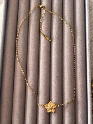Guldhalsband med blomma - Snyggt halsband i guldfärgad metall med en detaljrik blomma i mitten. Kedjan är tunn och har ett justerbart lås för perfekt passform. Blomman har fem blad och ger en elegant och trendig look.