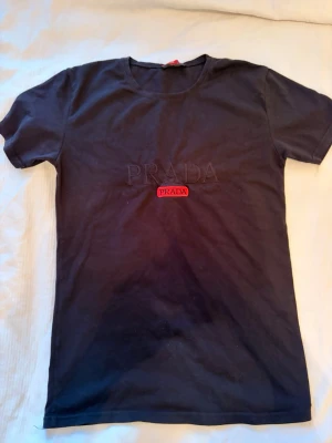 Svart t-shirt med PRADA-logga - Snygg svart t-shirt från PRADA med diskret broderad logga på bröstet och en röd PRADA-detalj. Klassisk rund halsringning och korta ärmar. Perfekt för dig som gillar stilrena och exklusiva plagg.
