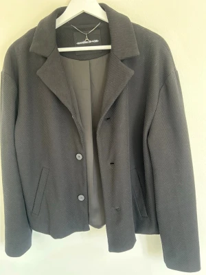 Svart jacka från Manière De Voir - Stilren svart jacka  från Manière De Voir med klassisk krage. Jackan har en diskret struktur i tyget och är helfodrad för extra komfort. Perfekt för dig som vill ha en clean och modern look. Max använd 5 gånger och mycket bra skick.