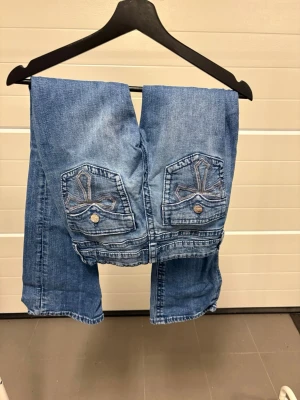 Blå jeans med broderade bakfickor - Säljer ett par blå jeans i klassisk tvätt med bootcut och snygga broderade detaljer på bakfickorna. Jeansen har knappar och fickor både fram och bak.