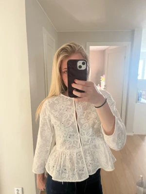 Vit broderad blus med volang - Säljer en vit blus med broderade blommor och hålmönster. Blusen har rund halsringning, markerad midja med volang och långa ärmar. 