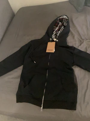 Svart hoodie med dragkedja Burberry - Snygg svart hoodie från Burberry med dragkedja framtill och klassiskt rutigt foder i huvan. Hoodien har fickor på sidorna och justerbar dragsko i huvan. Perfekt för en stilren och avslappnad look.