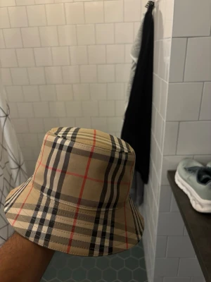 Burberry beige bucket hat - Säljer en klassisk bucket hat från Burberry i beige med det ikoniska rutmönstret i svart, vitt och rött. Hatten är tillverkad i bomull och har en mjuk, bekväm passform. Perfekt accessoar för att lyfta din stil med en tidlös touch.