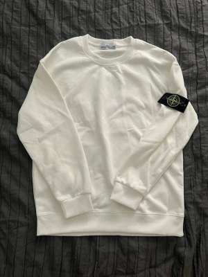 Stone Island långarmad. - Säljer en helt ny clean vit sweatshirt från Stone Island med den klassiska svarta patchen på ärmen. Tröjan har rund hals, ribbade muddar och är tillverkad i mjukt bomullsmaterial. Perfekt för dig som gillar streetwear och stilrena plagg.