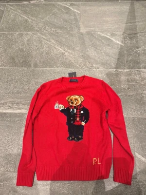  Polo Ralph Lauren - Röd stickad tröja från Polo Ralph Lauren med ikonisk björn i kostym och halsduk på framsidan. Tröjan har rund halsringning, ribbade muddar och RL-broderi nertill. Perfekt för dig som vill ha en unik och lekfull look.