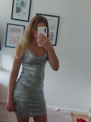 Kortklänning silver glittrig  - Snygg kort klänning i silver med glittriga paljetter och smala axelband. Klänningen har en figurnära passform och en lätt draperad urringning. Perfekt för dig som vill sticka ut och glänsa på nästa fest.