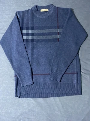 Mörkblå stickad burberry  - Snygg mörkblå stickad tröja med diskret rutmönster i grått och vinrött på framsidan. Tröjan har rund halsringning, långa ärmar och ribbade muddar vid ärmslut och nederkant. Perfekt för dig som gillar klassisk och stilren design. Storlek L skick ny 