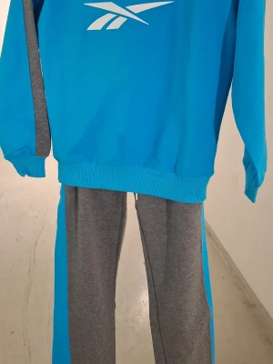 Blått och grått loungewear-set Reebok - Säljer ett snyggt loungewear-set från Reebok i blått och grått. Setet består av en sweatshirt med Reebok-logga och matchande byxor med dragsko och logga på benet. Perfekt för chill dagar hemma eller när du vill vara bekväm men ändå se fräsch ut.