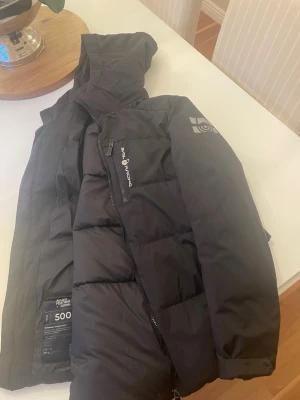 Svart vinterjacka Sail Racing med huva - Säljer en svart dunjacka från Sail Racing med huva och dragkedja. Jackan har logga på ärmen och bröstet samt en bröstficka med dragkedja. Fylld med dun och fjäder, perfekt för kalla vinterdagar. Snygg, sportig design och skön passform.