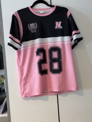 Rosa och svart t-shirt med tryck - Sportig t-shirt i rosa, svart och vitt med stora siffran 28 framtill och coola college-inspirerade detaljer. Kortärmad modell med rund hals och kontrastfärgade ränder på ärmarna. Perfekt för en avslappnad streetstyle-look.