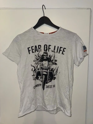 Vit t-shirt med motorcykeltryck - Säljer en vit t-shirt med trycket 'FEAR OF LIFE' och en cool motorcykelgrafik på framsidan. T-shirten har korta ärmar och ett amerikanskt flaggtryck på ena ärmen. 