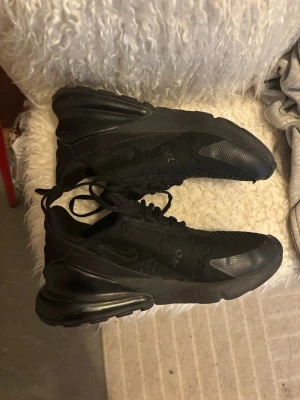 Svarta Nike Air Max 270 sneakers - Säljer ett par helsvarta Nike Air Max 270 sneakers med mesh-ovandel och synlig Air-enhet i hälen. Skorna har en sportig look med snörning och diskreta loggor på sidan och plösen. Perfekta för dig som gillar stilrena och bekväma sneakers.