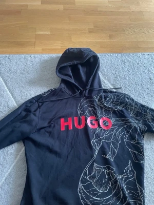 Svart hoodie från HUGO med tryck - Snygg svart hoodie från HUGO med stort rött logotryck på bröstet och vita grafiska linjer som motiv på ena sidan. Klassisk huva med dragsko och mjukt material som känns skönt mot huden. Perfekt för en avslappnad streetwear-look.