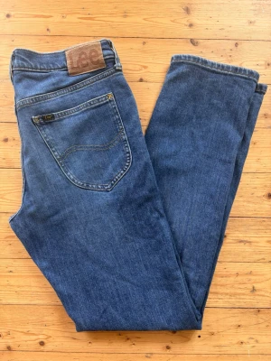 Blå raka jeans från Lee - Klassiska blå jeans från Lee med rak passform och fem fickor. Jeansen har en snygg mellanblå tvätt. Tillverkade i slitstark denim med gul kontrastsöm. Perfekta för en avslappnad och tidlös stil.
