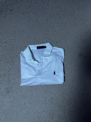 Vit pikétröja från Polo Ralph Lauren - Klassisk vit pikétröja från Polo Ralph Lauren med krage, knappar framtill och den ikoniska loggan broderad på bröstet. Tröjan har korta ärmar och är tillverkad i mjuk bomull som känns skön mot huden. Perfekt till jeans eller shorts för en clean look.