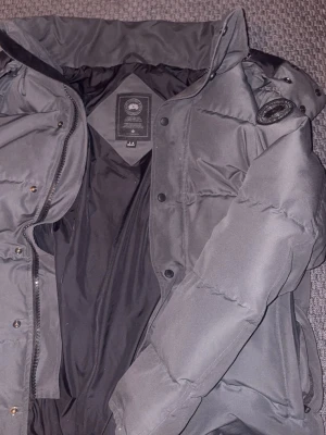 Svart dunjacka med patch på ärmen - Säljer en svart dunjacka med hög krage och dragkedja samt tryckknappar. Jackan har en patch på vänster ärm och quiltad design som ger en schysst streetlook. Perfekt för kalla vinterdagar tack vare det tjocka fodret.