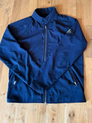 Stone Island windbreaker - Snygg marinblå vindjacka från Stone Island med klassisk patch på ärmen. Jackan har hög krage, dragkedja framtill och två snedställda fickor med dragkedja. Materialet är tunt och smidigt, perfekt för blåsiga dagar.