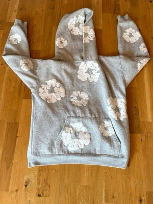 Denim tears Hoodie - Säljer en grå hoodie från Denim Tears i storlek S. Hoodien har vita blommönster över hela plagget och en stor ficka framtill. Tillverkad i mjukt material och har en klassisk huva. Perfekt för dig som vill sticka ut med en unik design.